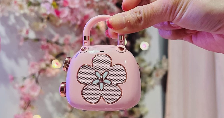 hand holding a up a mini Bluetooth speaker shaped like a purse, pink flowers in the background