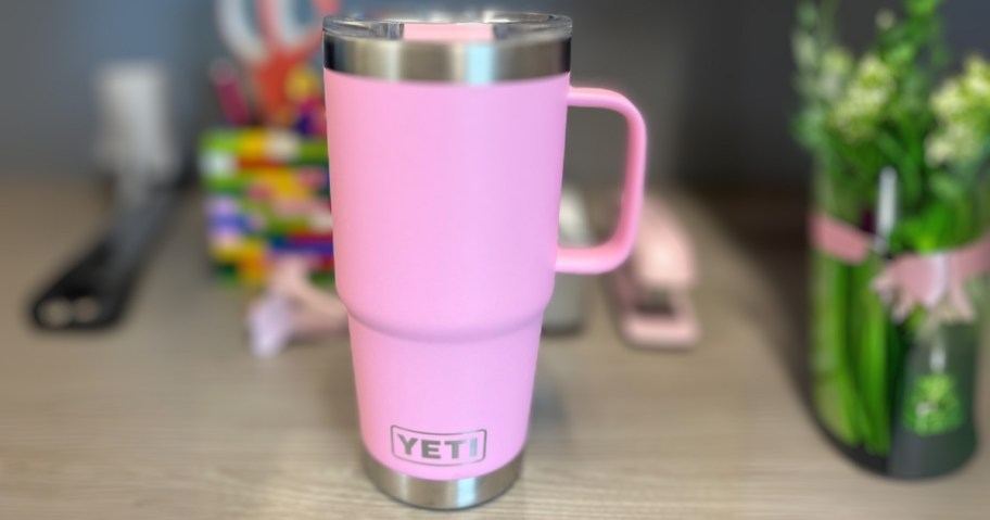 a pink YETI tumbler with a handle on a desk, offer items behind it and a vase of flowers to the side