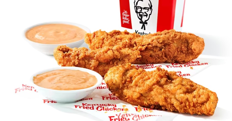 3 KFC chicken tenders on a KFC paper wrapper next to small bowls of sauce, KFC fries box in the background