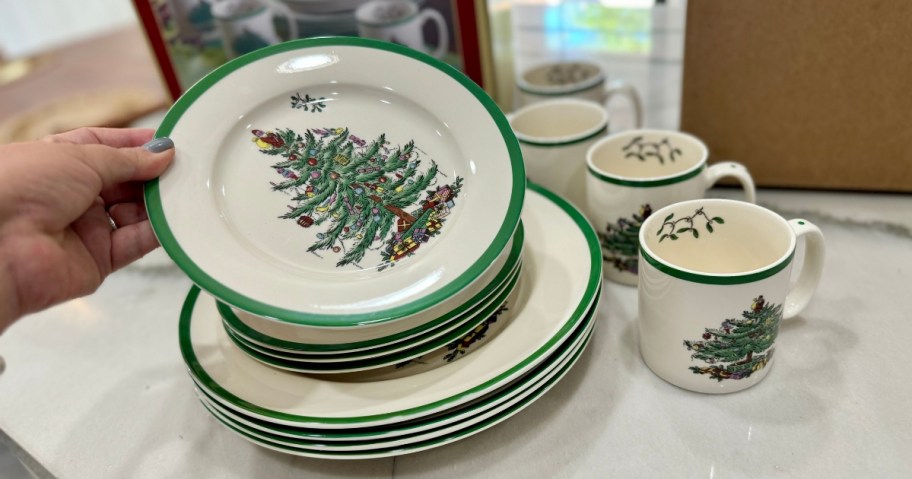 hand reaching to pick up a set of dinnerware that has Christmas trees on it, more plates and matching mugs on the table around it