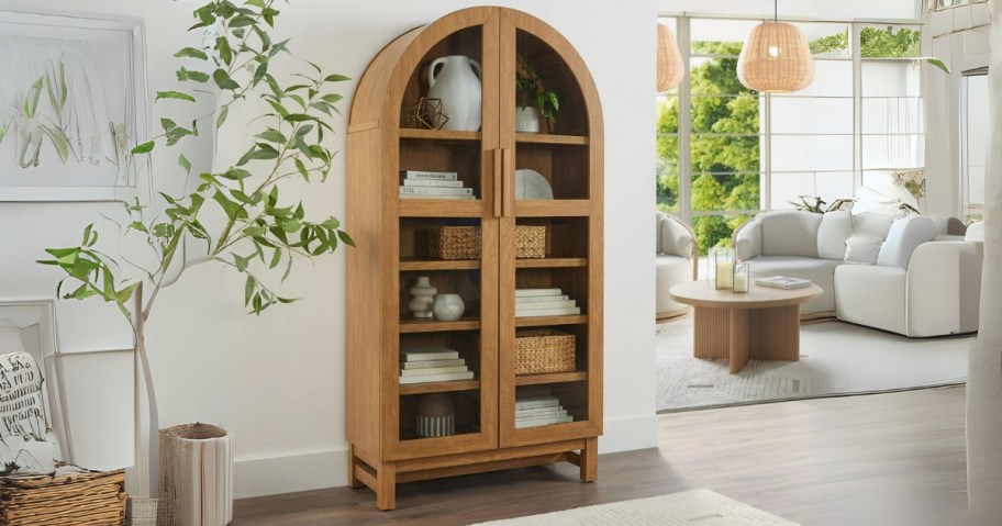 a light oak wood arched cabinet with glass doors in a hallway, living room off to the side