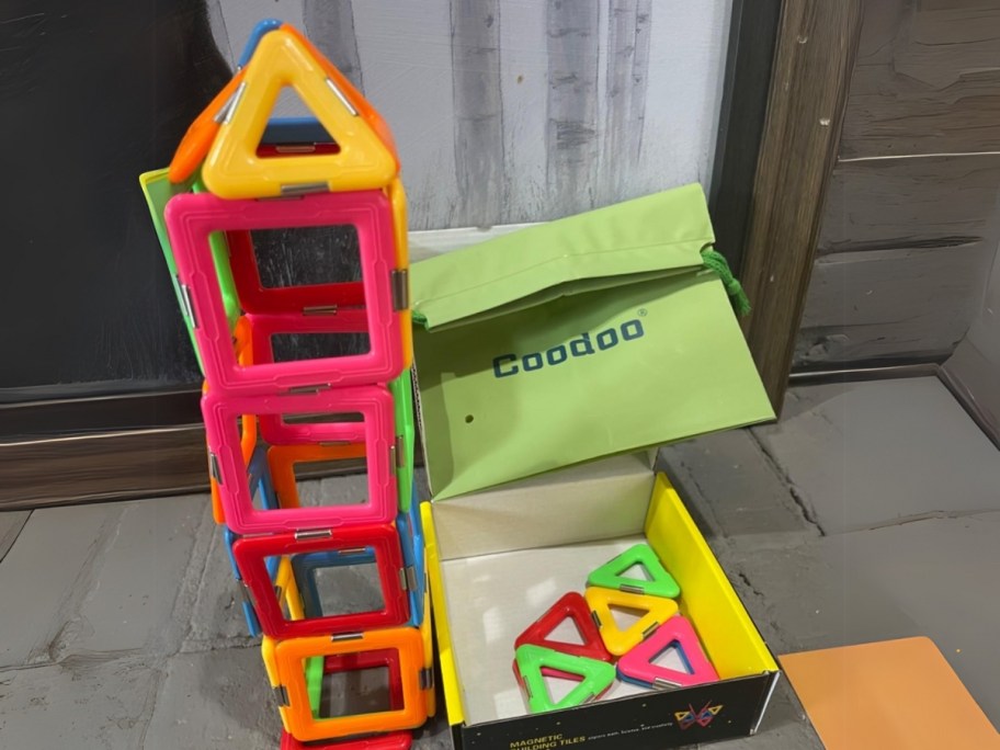 a set of colorful magnetic building tiles shaped like a house, the box they onlinee in and a green storage bag sits to the side