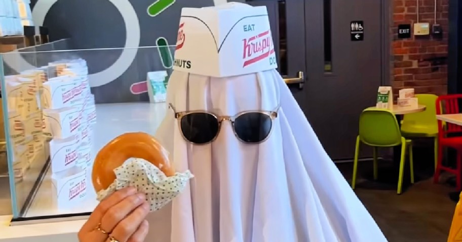 person wearing ghost halloween costume and holding glazed doughnut