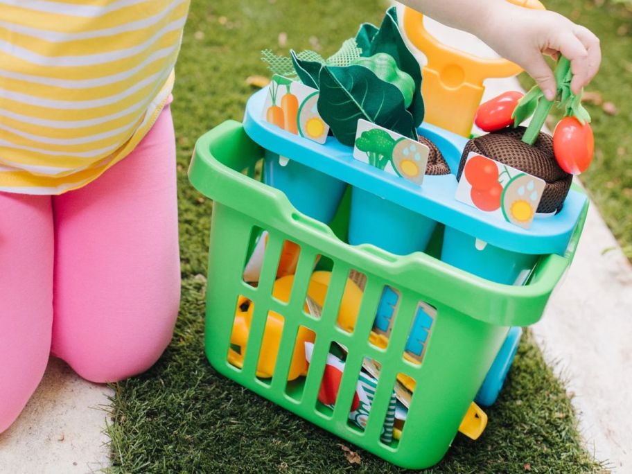 girl sitting next to Melissa & Doug Let's Explore Vegetable Garden Play Set w/ Plastic Rolling Cart outside