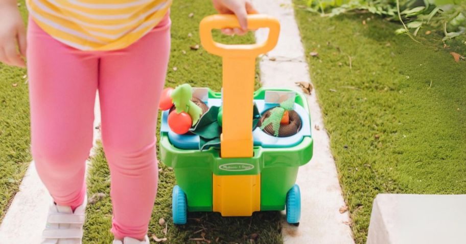 girl pulling Melissa & Doug Let's Explore Vegetable Garden Play Set w/ Plastic Rolling Cart outside