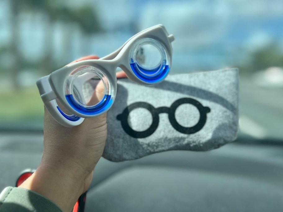 hand holding a pair of white and blue motion sickness glasses and a grey zipper storage pouch for them, car's dash and front windshield behind them