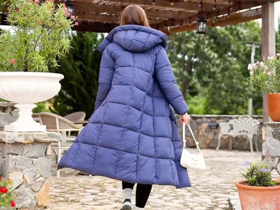 A woman in a long navy blue puffer coat standing with her back to us in a garden