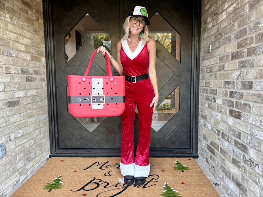 Woman standing in front doorway outside wearing a Santa jumpsuit holding a Santa bogg bag 