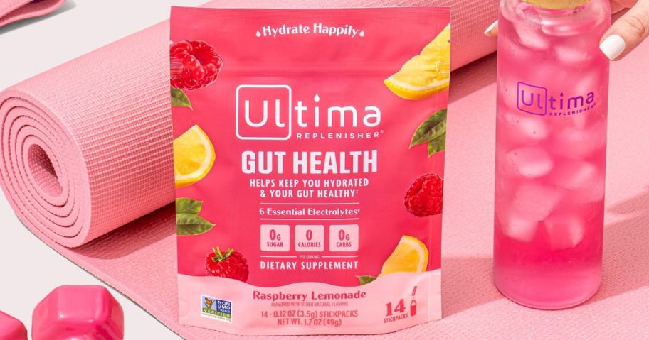 A bag of Ultima Replenisher Gut Health Drink Mix next to a glass water bottle filled with a pink liquid