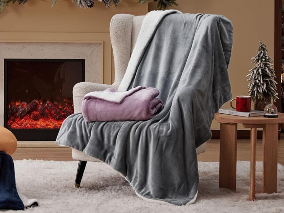 an accent chair with a grey sherpa fleece throw blanket spread over it and a light purple blanket folded on it, fireplace in the background