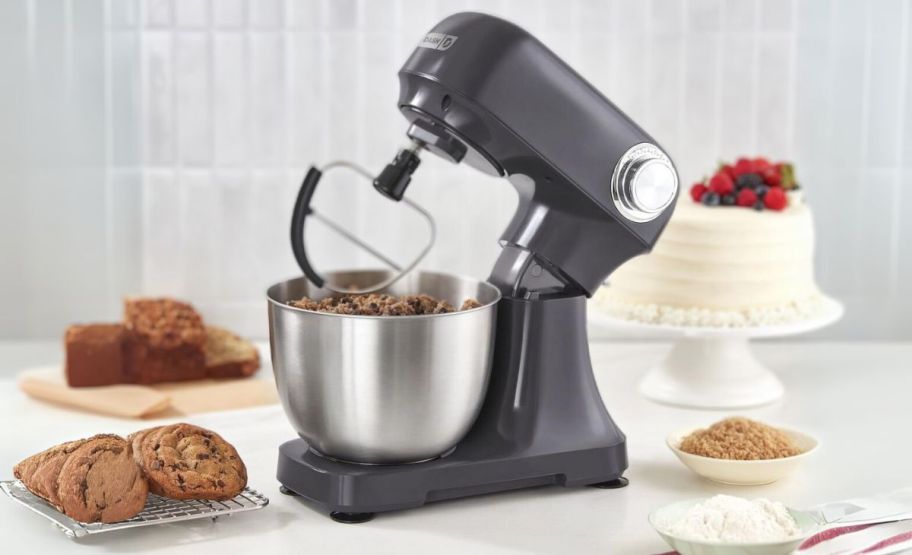 a gray dash stand mixer with head tilted, shown with cakes, cookies, and breads