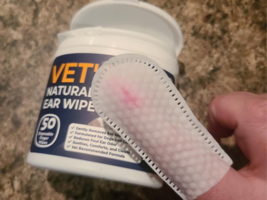 finger with a pet ear wipe on it, the container it came from behind the persons hand