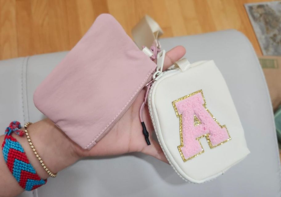 A womans hand holding a [ink and beige dual pouch wristlet with monogram letter