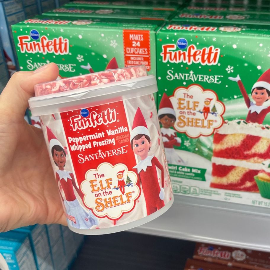 a womans hand holding a tub of elf on a shelf funfetti frosting