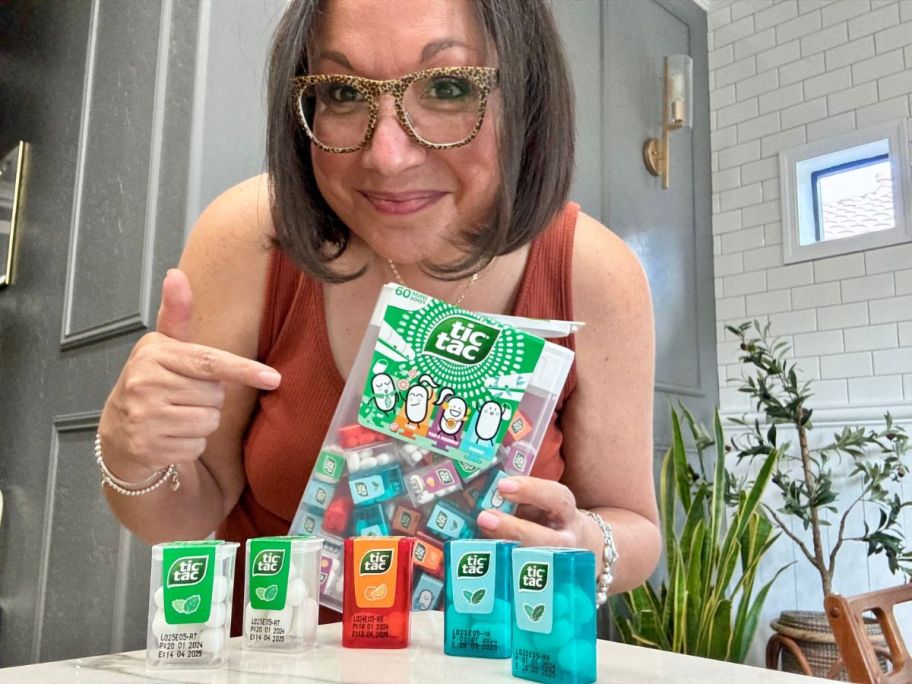 woman with brown hair and glasses smiling at the camera holding a giant Tic Tac candies box with mini Tic Tac containers inside, and some of them on a counter in front of her