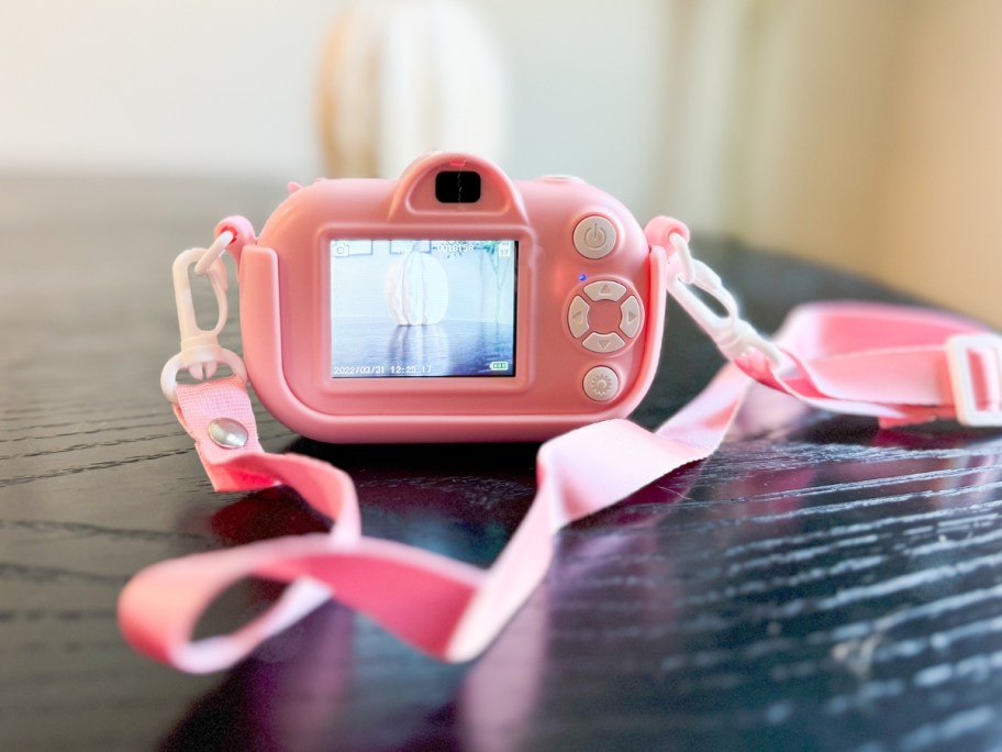 the back of a kid's pink digital camera showing the screen, the camera has an attached lanyard and is sitting on a table