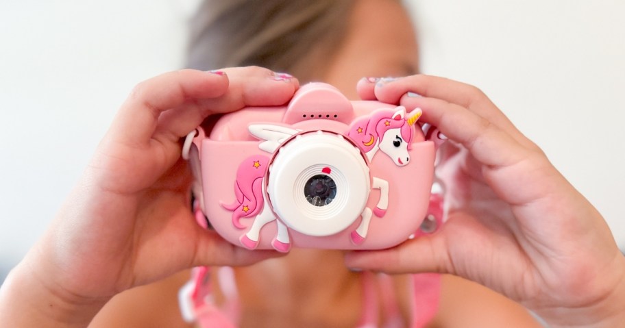 little girl holding a kid's camera up and taking a picture with it, the camera is pink with a unicorn on it and has a lanyard strap