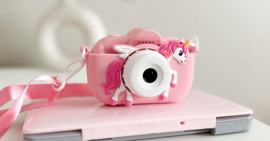 a kids digital camera in pink with a unicorn on it and an attached lanyard on top of a kids laptop