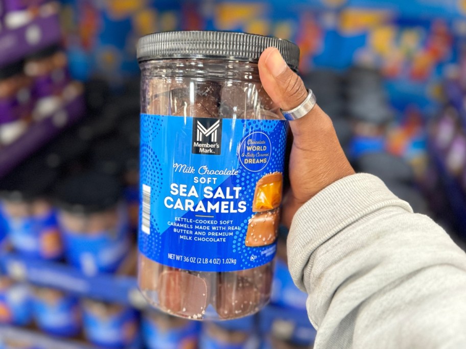 hand holding a jar with a blue label with Member's Mark Milk Chocolate Sea Salt Caramels in it, more on a shelf behind it