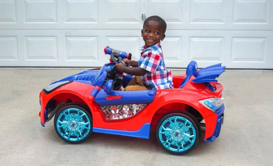 a little boy driving a spider man super car ride on in a driveway