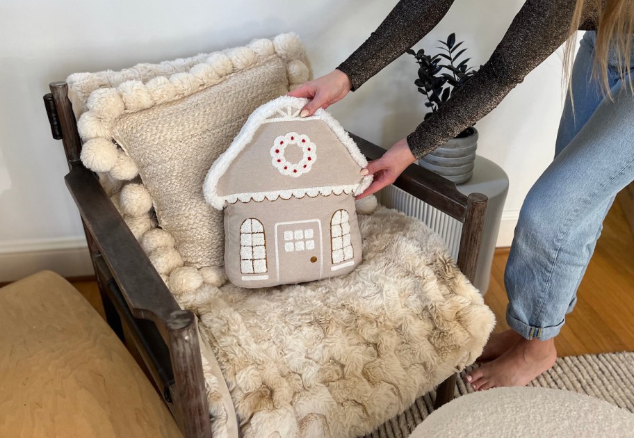 woman placing neutral gingerbread house pillow on chair with fur blanket