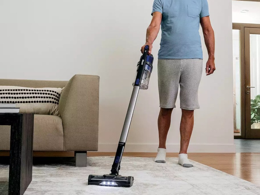 man using a blue cordless stick Shark vacuum to vacuum the floor in a living room