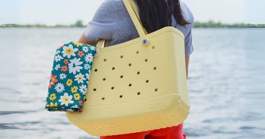 woman facing the ocean wearing a blue shirt and red shorts and yellowing a large yellow Bogg bag with a green floral accessory bag