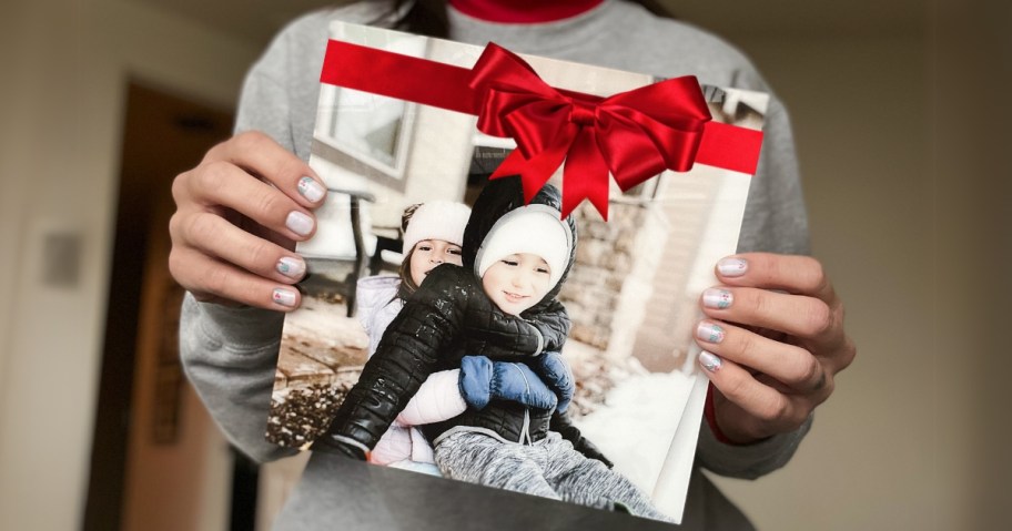 a woman holding a photo in a glass frame with no border of 2 kids in winter coats playing outside, there is a red bow at the top of the photo