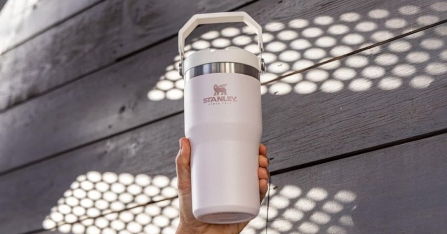 hand holding a Stanley IceFlow Tumbler in a light pink Rose Quartz color, wood wall behind it