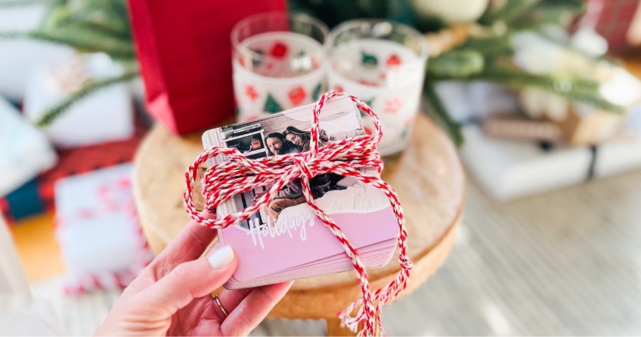 hand holding a stack of photo coasters with ribbon tied around them, holiday candles and gift back on a stool behind it