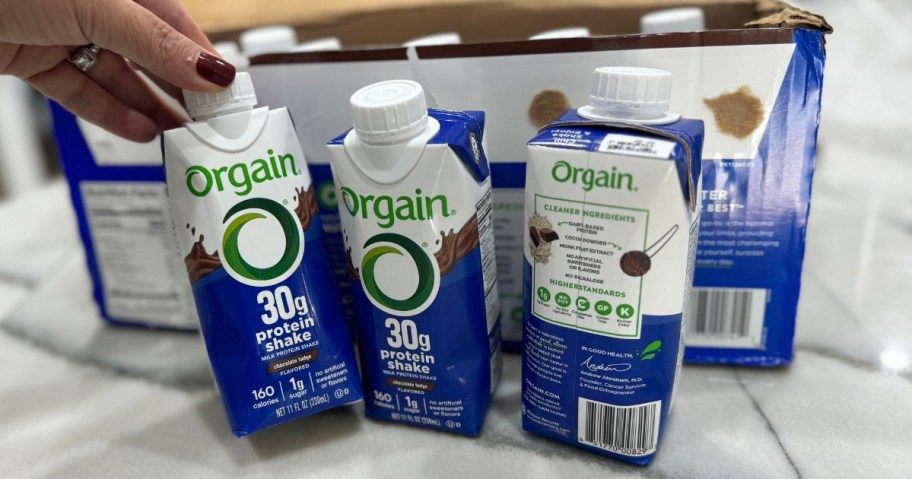 a hand reaching for a ready to drink Orgain shake bottle, 2 more beside it and box that a pack of 12 onlinees in behind it