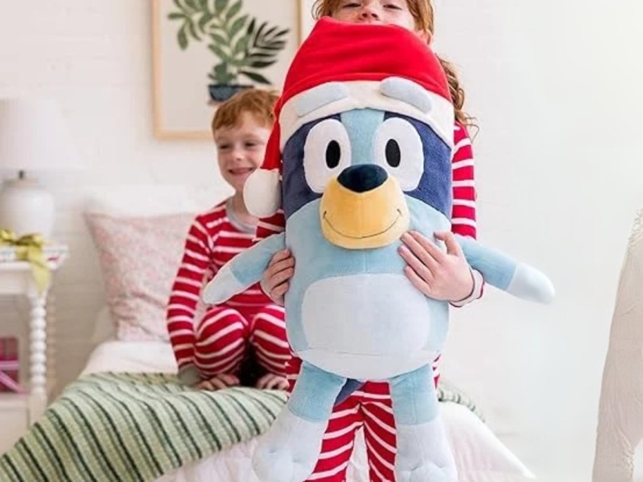 kid holding a jumbo 24 inch christmas holiday Bluey plush in a Santa hat, little boy on the bed behind her