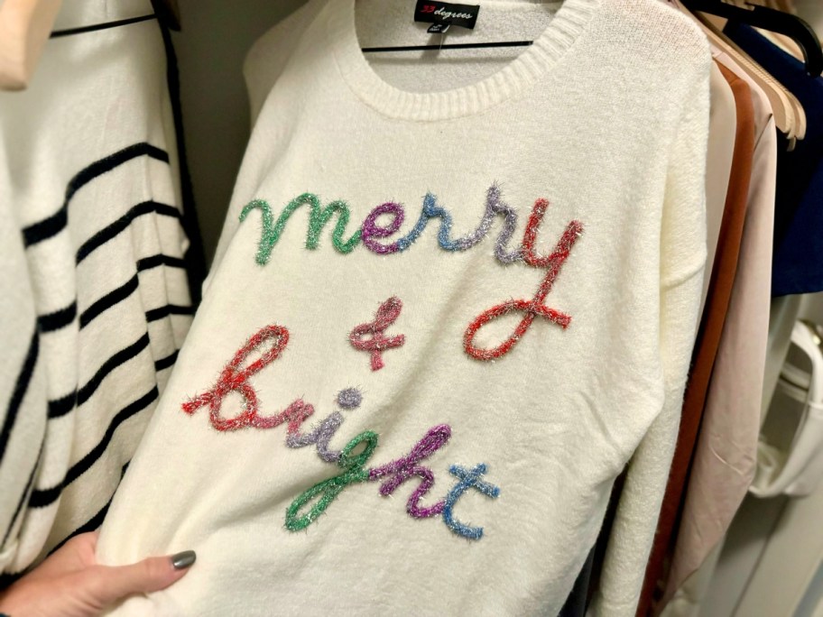 woman's hand pulling a sweater out of a closet that's on hanger. The sweater is a cream color and says "merry & bright" in colorful embroidered letters