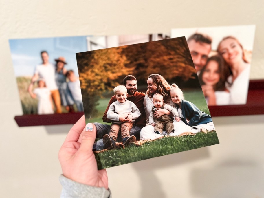 woman holding a glass framed photo of her family with no border on the frame, wall shelf with more glass framed family photos behind it
