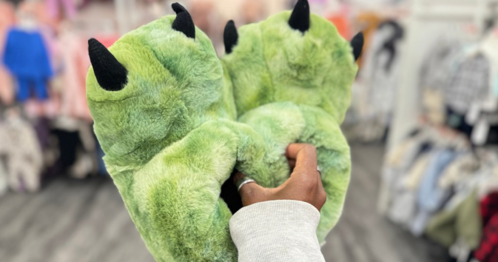 person holding green dinosaur slippers in target