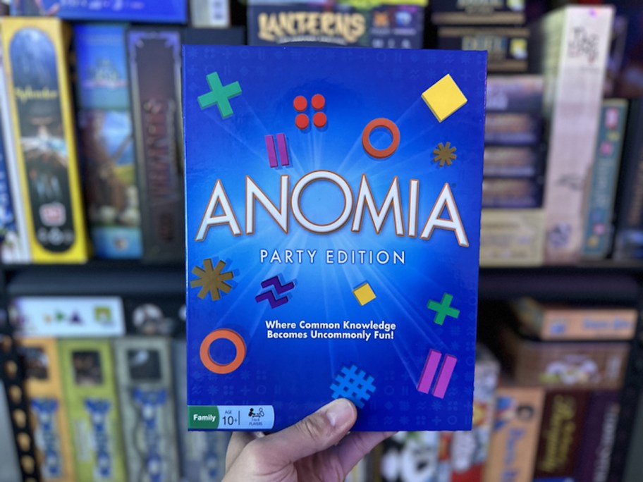 hand holding anomia board game in front of shelf