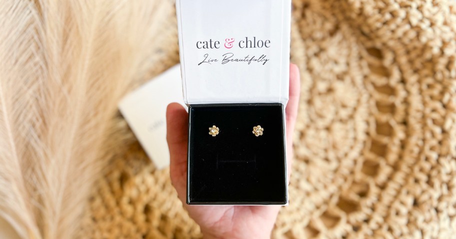 hand holding cate and chloe earrings in box over straw hat 