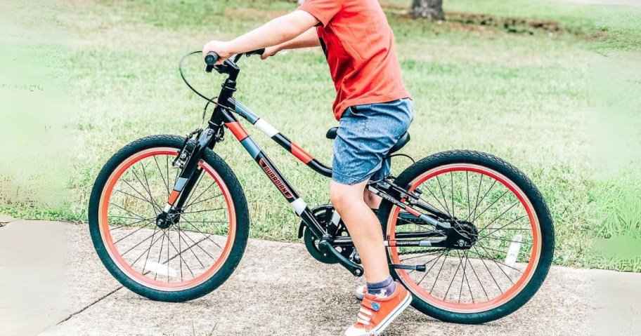 little boy wearing an orange top and jean shorts and orange Chuck Taylor shoes riding on a black and orange Guardian bicycle on a sidewalk