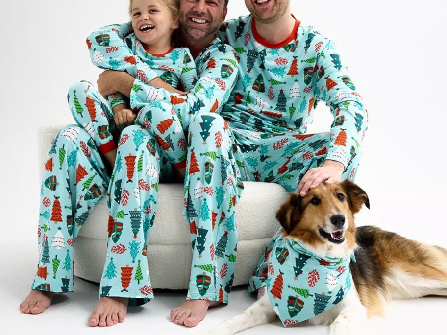 two dads and a little girl and a dog wearing matching family Christmas pajamas that are blue and have multi color Christmas trees on them, the dog has a matching bandana on