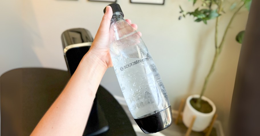 hand holding a clear and black SodaStream bottle, SodaStream machine on table behind it