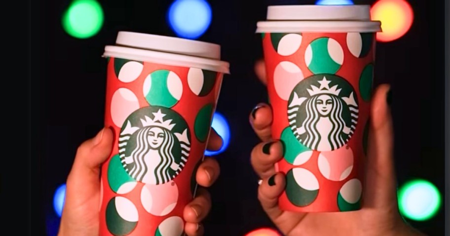 two hands holding Starbucks to go cups with red, green, pink and white holiday designs, Christmas lights behind them