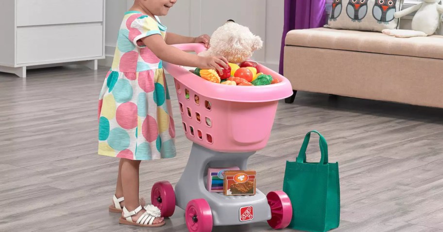 toddler putting a stuffed bear into a pink step 2 shopping cart full of pretend food