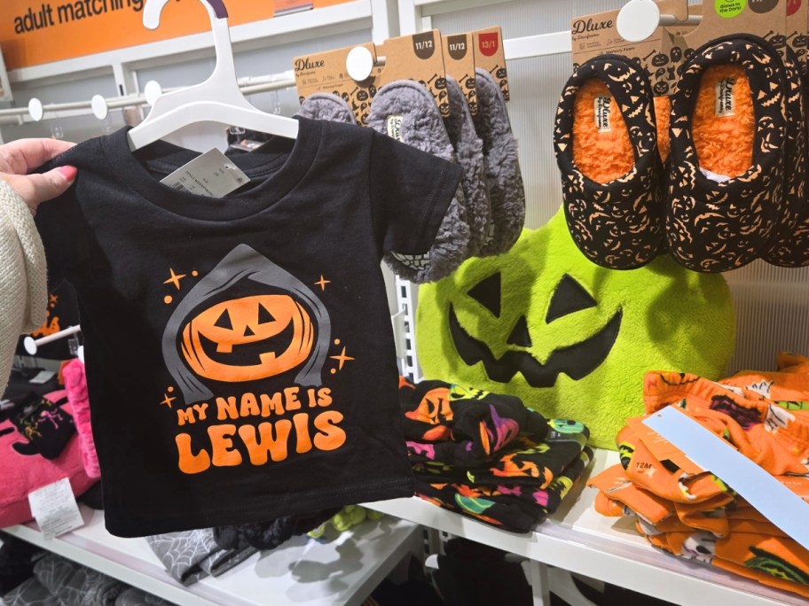 hand holding up a kid's halloween tshirt, halloween slippers and more behind it