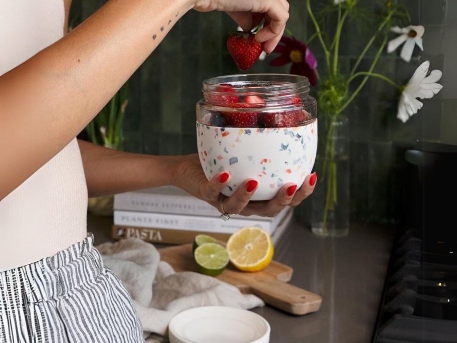 woman holding a glass food bowl with a cream and terrazzo print silicone sleeve, she is pulling a strawberry out of it, kitchen counter is behind her and you can see a cutting board, lemon slices and the lid for the food container