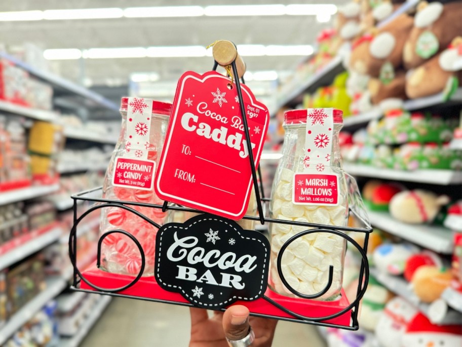 hand holding a black metal caddy with glass bottles in it and a black and white sigh that says "Cocoa Bar" on it