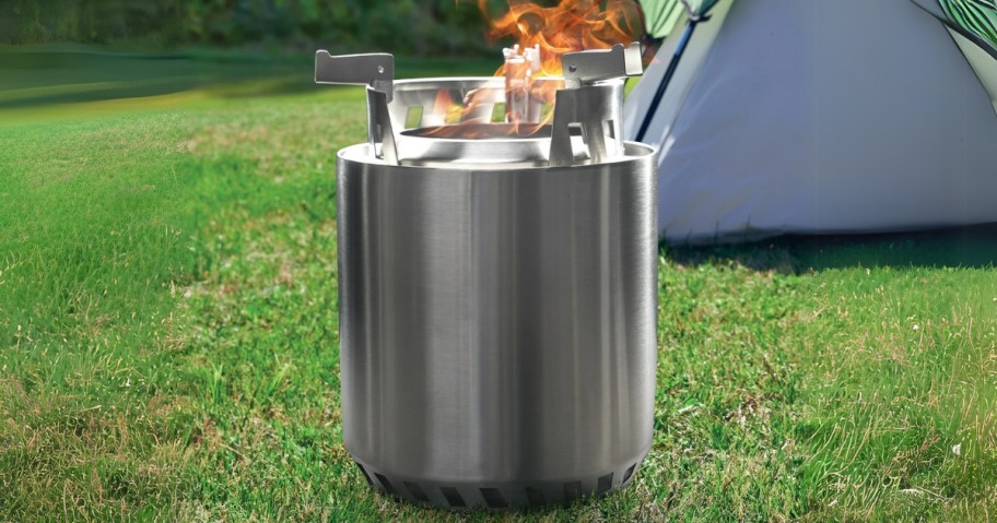 a silver metal camp wood burning fire pit outdoors, tent in the background