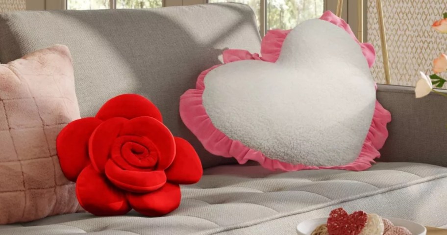 a grey sofa with a red rose shaped pillow and a large white heart shaped pillow with a pink ruffle