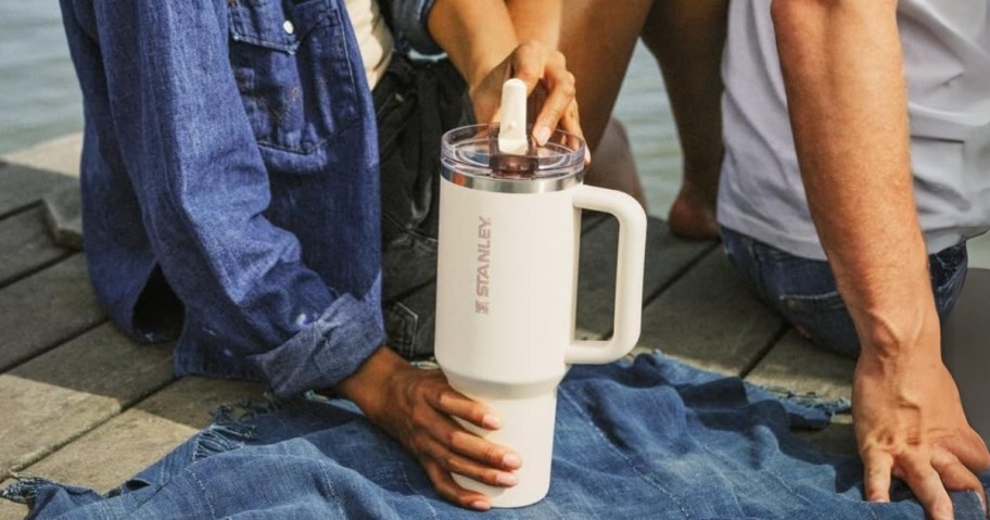 woman and man sitting on a dock by a lake, woman is holding a cream color Stanley tumbler with a flip straw lid and handle