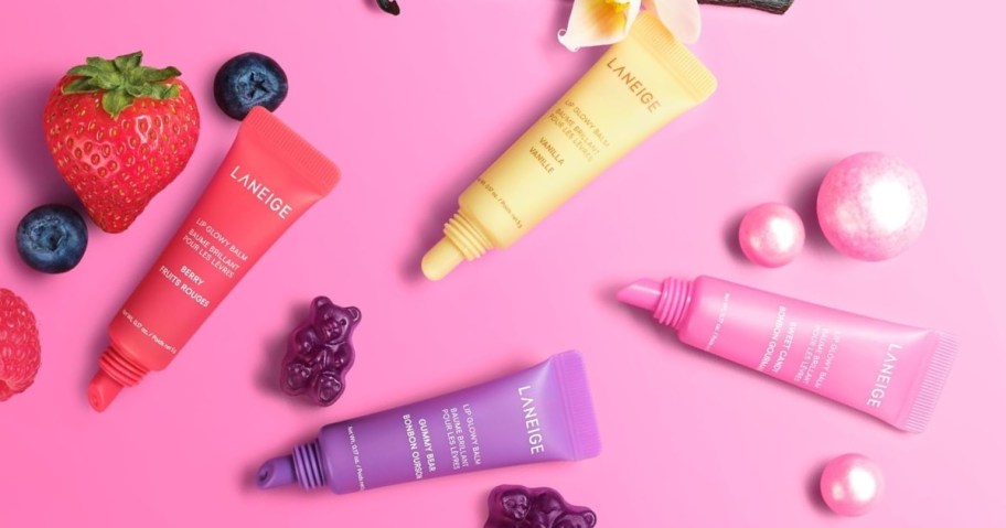 a 4 piece set of Laneige glowy lip balms in dark pink, purple, yellow, and light pink colors on a pink background surrounded by fruit