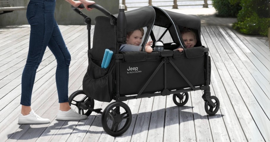 a woman pushing a stroller wagon that has a double canopy up, you can see 2 small kids inside